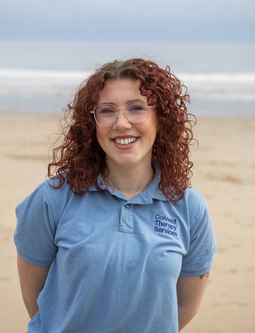 Ellie Nichol with curly red hair and glasses, smiling on the beach in a light blue Connect polo shirt