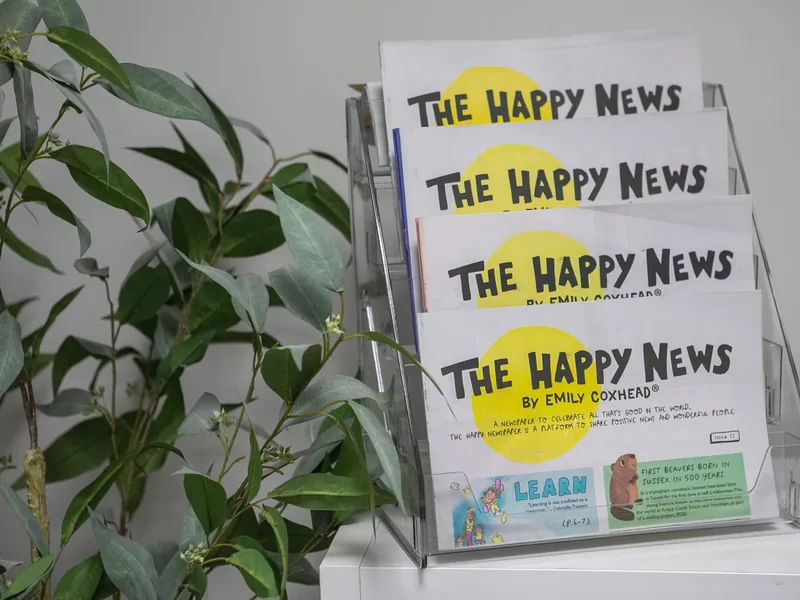 Copies of The Happy News newspaper in a display rack beside a plant in the Connect clinic