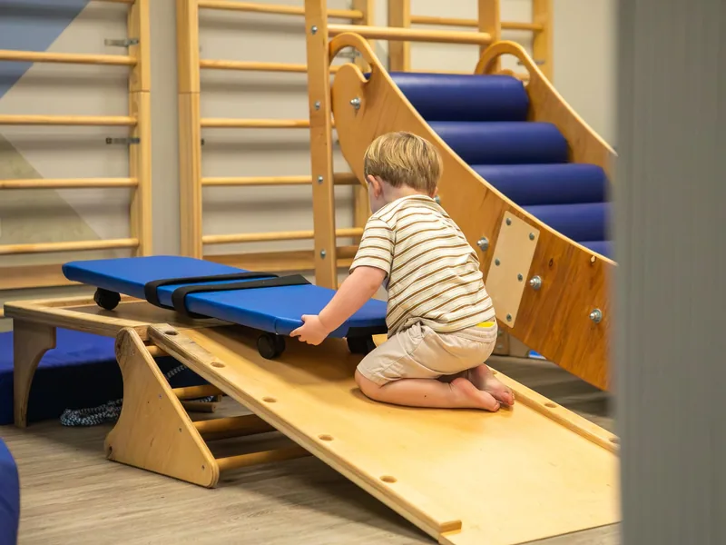 A young boy kneeling on a wooden ramp pushing a scooter board in the Connect soft play room