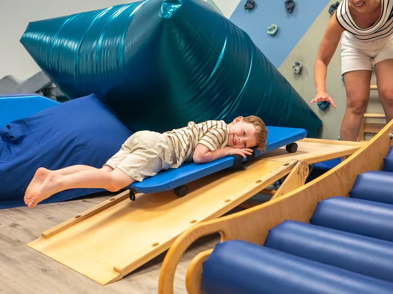 A smiling young boy lying on a scooter board on a ramp while a Connect therapist watches