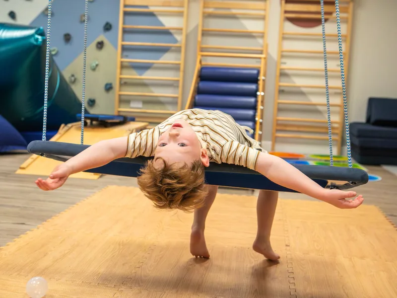 A young boy draped upside down over a therapy swing with his arms outstretched