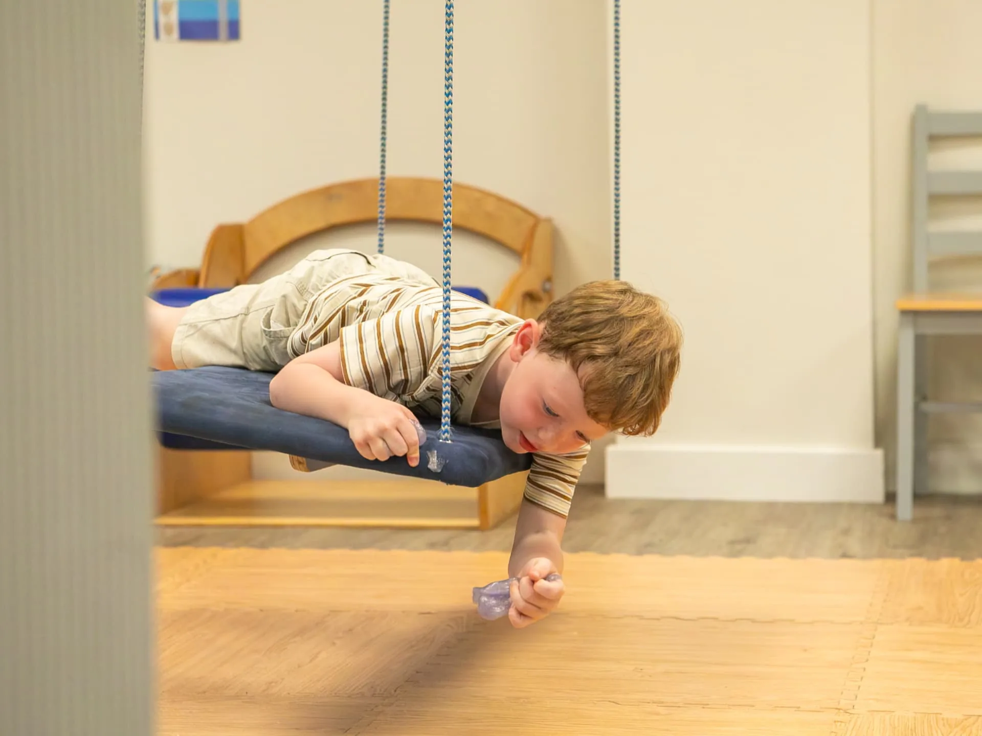 A young boy lying on his tummy on a therapy swing, reaching for the floor